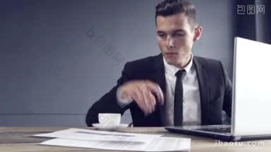 英俊的年轻商人在笔记本电脑上工作，喝着咖啡，对工作结果感到满意