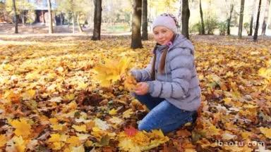 可爱的少女坐在她的膝盖上，收集从树上飘落的黄叶在美丽的秋天城市公园
