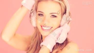 快乐的微笑的金发女人在粉红色的冬季手套和耳罩在粉红色的工作室背景