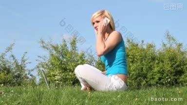 漂亮的金发女人坐在郁郁葱葱的绿色草坪上，她的笔记本电脑在她的前面，在她的<strong>手机</strong>上说话