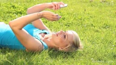 年轻漂亮的金发女孩躺在她的绿色草地上阅读短信在<strong>手机</strong>上