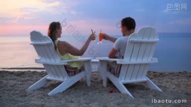 一对浪漫的情侣坐在沙滩上的木质躺椅上，一起举杯庆祝日落，在鸡尾酒<strong>杯中</strong>碰杯，从背后眺望风景