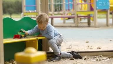 在户外的儿童游乐场里，一个小男孩蹲在一张五颜六色的小桌子前玩玩具拖拉机