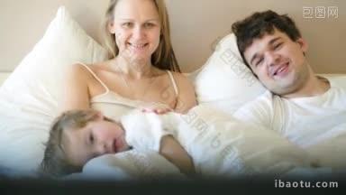 年轻的父母和儿子早上躺在床上看电视，父亲拿着遥控器换频道