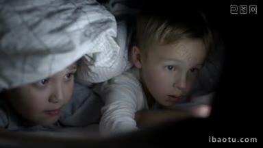 两个男孩晚上躺在床上的毯子下用触摸板看动画片或电影