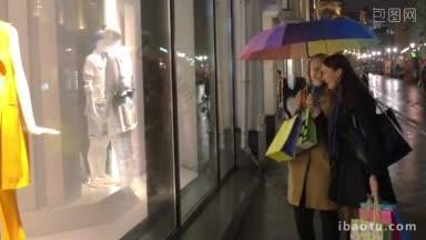 年轻的妇女在购物的晚上，他们拿着雨伞和购物袋，看着橱窗和讨论展示的<strong>衣服</strong>