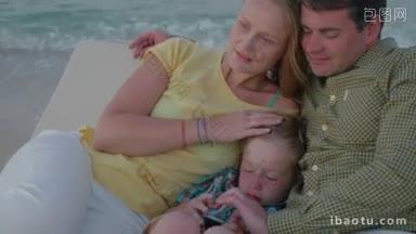 年轻的家庭在躺椅上放松，在海边聊天，父亲和儿子玩手机，而母亲