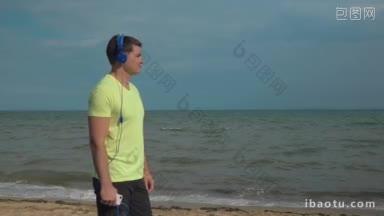 一个年轻人站在海滩上，用智能手机戴着耳机听音乐的慢动作自然场景