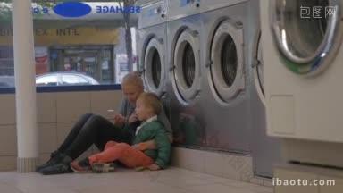 母亲和儿子在洗衣房等待，他们坐在洗衣机附近的地板上使用智能手机