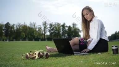 年轻的<strong>女</strong>商人赤脚坐在公园的草地上，在户外用笔记本电脑工作
