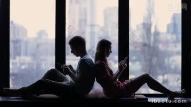 剪影的年轻夫妇坐在家里背靠背和使用智能手机的窗口背景上网上瘾的夫妇使用