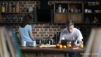 英俊的潮人在笔记本电脑上浏览社交媒体，而他的女朋友在厨房里准备早餐，年轻夫妇在一起休闲