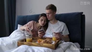 年轻迷人的情侣穿着睡衣，用智能手机在床上看视频<strong>内容</strong>，在早上享受美味的早餐