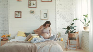 快乐的准妈妈。采取自拍照片怀孕了女人坐着床上