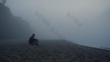 抑郁的家伙坐着海海滩多雾的早....担心男人。触碰沙子