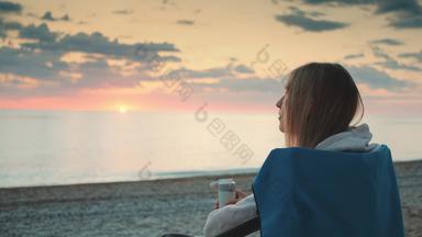 年轻的女人<strong>喝热水</strong>瓶坐着野营椅子海滩
