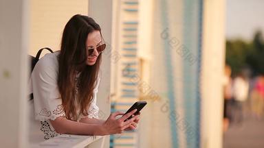 年轻的高加索人女人发送消息智能手机户外公园日落美丽的女孩太阳镜坐着木板凳上智能手机