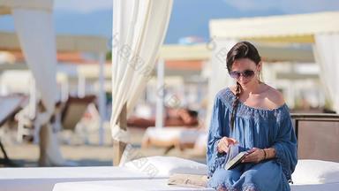 年轻的女人阅读书热带海滩假期时尚女孩读坐着白色日光浴浴床欧洲海滩周末
