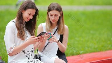 年轻的女孩聪明的电话在户外女性坐着公园咖啡智能手机