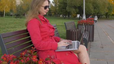 年轻的高加索人业务女人太阳镜红色的斗篷工作移动PC坐着板凳上城市公园红色的花前景<strong>替身</strong>移动拍摄帧/秒