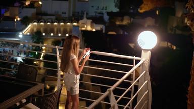 少年女孩持有移动电话晚上阳台年轻的女孩智能手机户外阳台照明灯笼背景度假胜地酒店