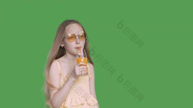 年轻的女孩喝橙色汁玻璃稻草绿色背景少年女孩喝水果鸡尾酒透明的绿色背景α通道键控绿色屏幕