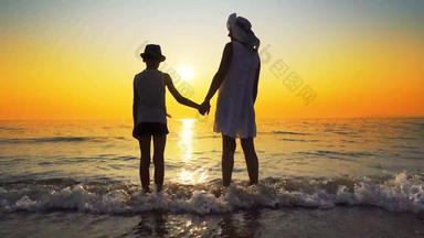 旅行家庭爱在一起概念妈妈。儿子穿帽子站空海滩持有手看令人惊异的日落电影替身拍摄