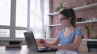<strong>在线</strong>培训，戴眼镜的英俊年轻女子使用笔记本电脑，并在坐在厨房办公桌前的剪贴板上写笔记