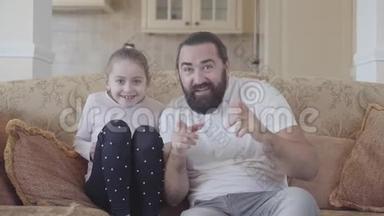 大胡子男人坐在沙发上看电视，他的小女儿支持足球队关门。 女孩跳起来