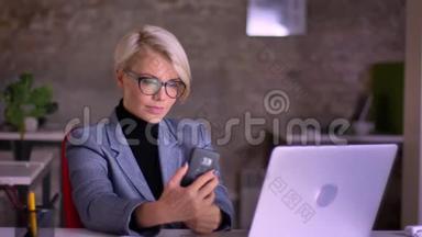戴眼镜的中年金发短发女商人在办公室用手机制作自拍照片的肖像。