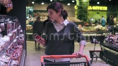 年轻的深色头发的女顾客在杂货店里穿过肉道时看着手机。 女孩带着腊肠