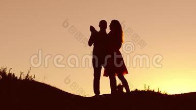 恋爱中的情侣在日落时跳舞剪影和接吻。 爱的男人和女人与狗跳舞剪影缓慢