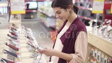 年轻漂亮的黑发女孩在电子商店摘电话时微笑。 查看智能手机应用程序