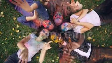 欢乐的女孩和男孩躺在公园的草地上，他们的脸和衣服上涂满了多色的油漆，人们都喜欢