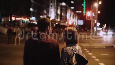 年轻时尚夫妇站在红绿灯前的后景。 美丽的男人和女人在晚上过马路。