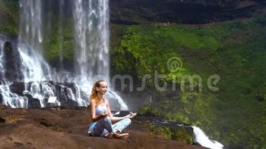 女孩在瀑布的大石头上摆着普拉纳亚摩瑜伽姿势