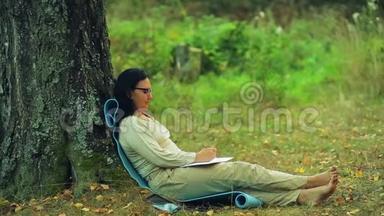 一个戴眼镜的女人光着脚<strong>坐在</strong>公园的一棵<strong>树下</strong>，在笔记本支铅笔