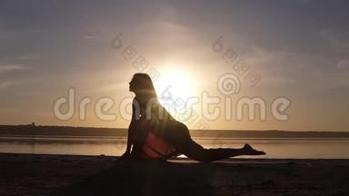 美丽的瑜伽女人的剪影在清晨的天空中在一个海边。 一个练习瑜伽的女人维拉巴德拉萨纳二世勇士