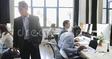 老板走在现代的开放式办公室，而一群商业人士在电脑上工作，领导看混合种族小组