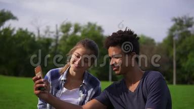 可爱的朋友在公园里休息，坐在草地上，看着智能手机，笑着。 一对混血儿