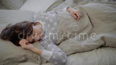一幅年轻的黑发女人的肖像，她穿着灰色睡衣，躺在床上，睡在家里用亚麻布覆盖的床上。 美丽美丽