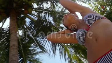 身着比基尼的年轻女子站在棕榈树下，太阳上晒着椰子