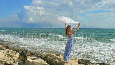 美丽的女士穿着长裙，在岩石海滩上挥舞着白色的围巾