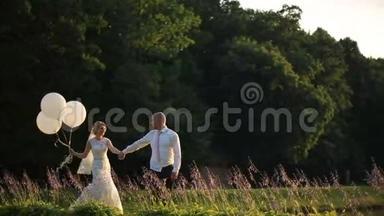 快乐的年轻新婚夫妇在夕阳下的夏日田野上带着气球散步。 浪漫婚礼概念