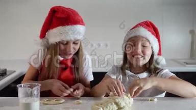 两个可爱的小女孩戴着圣诞老人的帽子，围着红色的圣诞围裙，亲手做饼干。微笑。新年快乐