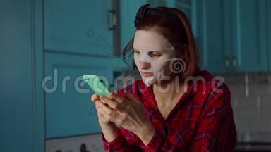年轻的30多岁的女人用手机在脸上戴着白色的化妆面具，在家里做自拍的鬼脸。 女人美丽