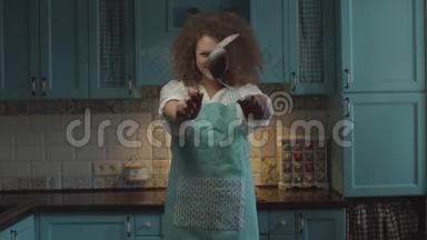 年轻卷发20多岁围裙女人手拿厨具站在蓝色厨房里，用搞笑的方式移动双手