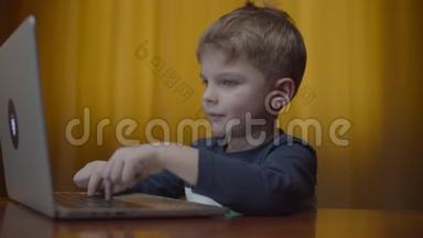 金发的学龄前男孩在笔记本电脑上打字，微笑着看着电脑屏幕。 孩子在家坐在网上学习
