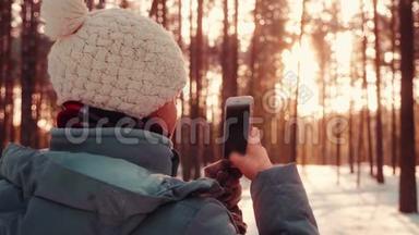 一位年轻漂亮的女士用手机在冬日的雪日落森林拍照
