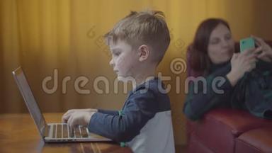 金发的学龄前男孩在笔记本电脑上打字，微笑着看着电脑屏幕。 妈妈拿着手机躺在沙发上